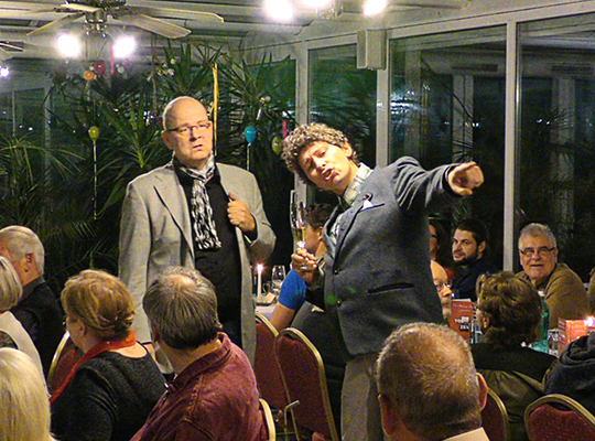 Zwei männliche Schauspieler stehen in der Mitte des Raumes und unterhalten das Publikum