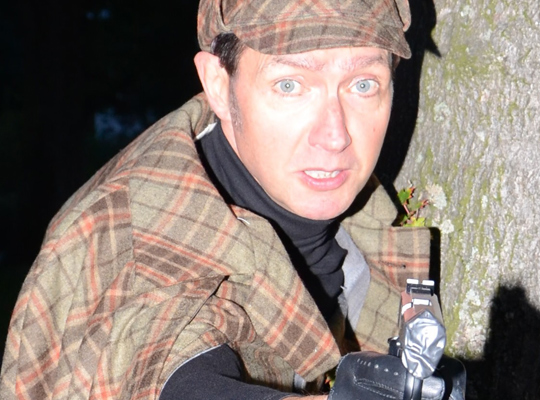 Angespannter Sherlock lehnt an einem Baum und zielt mit seiner Waffe auf eine Person.