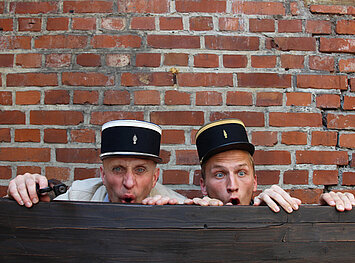 Hinter einem Holzzaun verstecken sich zwei Schauspieler mit Hüten, vor einer Steinmauer.