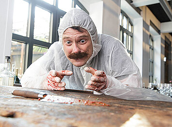 Fokussierter Blick auf ein Blutverschmierten Messers, welches auf einem Holztisch Liegt. Im Hintergrund Stehen leere Schnapsflaschen herum. Neben dem Messer liegt eine Zip Tüte.