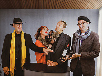 Eine Schauspielerin und drei Schauspieler stehen vor einer Wand an einem Stehtisch, die beiden äußeren unterhalten sich über einen teuren Wein, die Dame lehnt sich an den Herren und flirtet mit ihm.