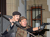 Dr. Watson versteckt sich hinter Sherlock Holmes, der eine Pistole zielend nach vorne hält.