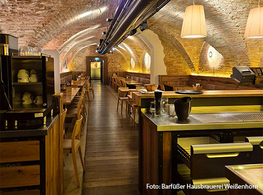 Langer Raum mit der Ansicht einer Bar aus, im Raum stehen hölzerne Tische und Stühle