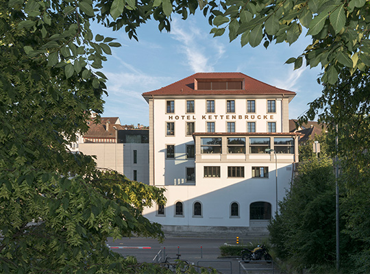 Blick von der gegenüberliegenden Straßenseite, durch zwei Bäume, auf das schöne Hotelgebäude