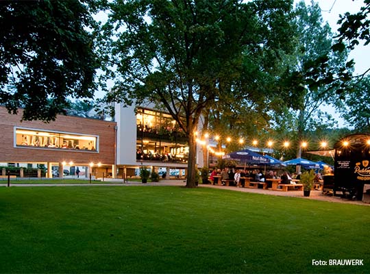 Große grüne Wiese vor einem Backsteingebäude, rechts neben dem Gebäude, befindet sich ein Biergarten