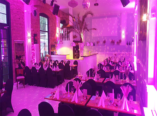 Ein Restaurant ist mit Stühlen und gedeckten Tischen ausgestattet. Lilanes Licht beleuchtet den Raum. 
