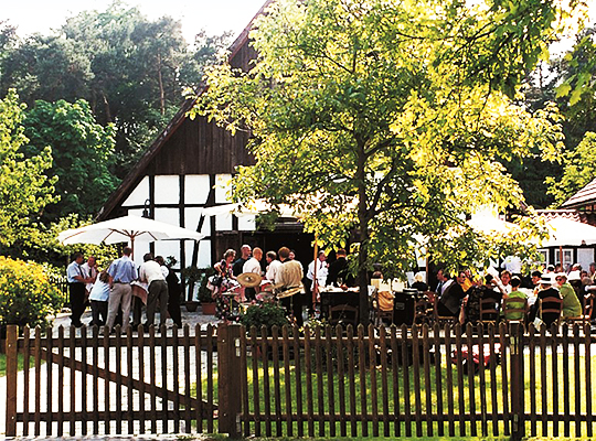 Blick auf das schöne Gelände des Gasthaus Buschkamp bei bestem Sonnenschein. Hinter einem braunen Holzzaun blüht alles schön grün.