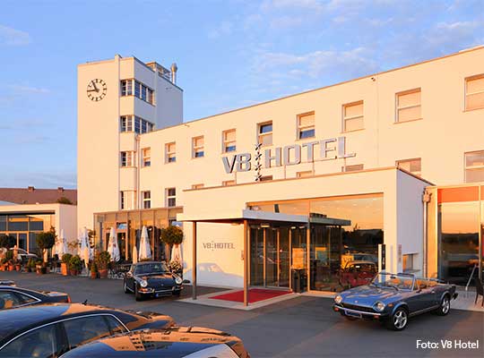 Sonnenbestrahltes V8 Hotel-Gebäude inklusive Parkplatz gefüllt mit Oldtimern.