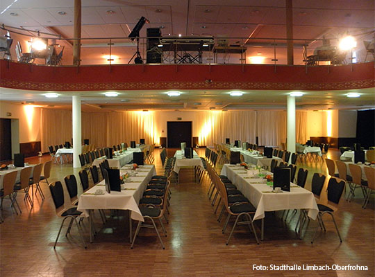 Innenansicht der Krimidinner in der Stadthalle Limbach-Oberfrohna mit langen Tischen, auf welchen Weiße Tischdecken liegen. Unter den Tischen stehen Stühle