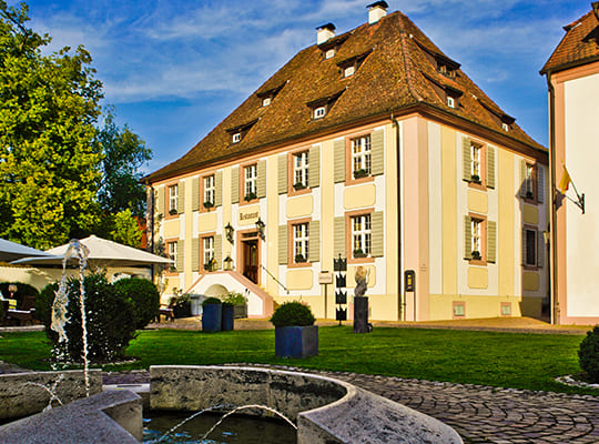 Blauer Himmel über dem von der Sonne bestrahlten Hotel-Gebäude inklusive Springbrunnen und Schlossgarten