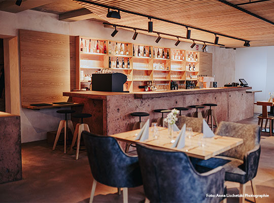 Innenraum mit Ansicht auf Bar mit kleinem Tisch an welchem vier Stühle stehen. Der Tisch ist mit Besteck, vier Servietten und einer Blume in Vase gedeckt.