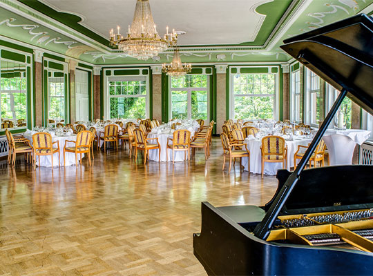 Ein festlicher Ballsaal mit einem schwarzen Piano, edel gedeckten Tischen und einer großen Tanzfläche 