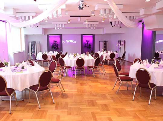 Krimidinner in großem Raum mit runden Tischen, Girlanden an der Decke und lila Beleuchtung