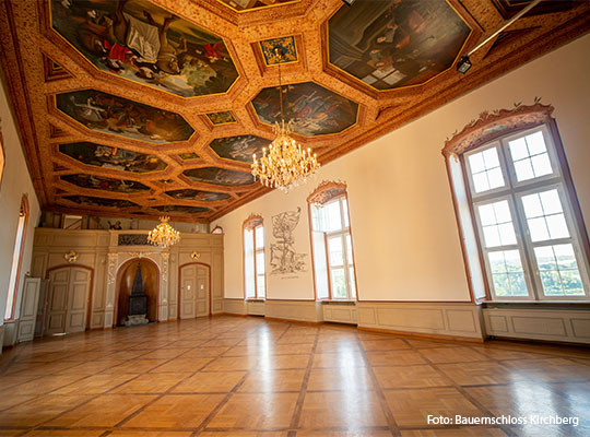 Innenansicht des Schlosses mit hoher verzierter Decke und ohne Stühle