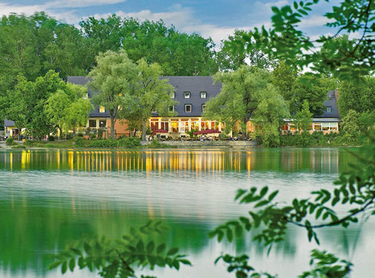 Wunderschöner See mit dem in Bäumen versteckten Dinnerkrimi Tatort-Hotel im Hintergrund, die Farben spiegeln sich idyllisch auf der Wasseroberfläche