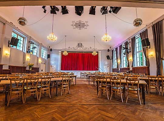 Schön geschmückter Saal mit dunklem Holzboden und großen Kronleuchtern an der Decke, die Tische und Stühle sind links und rechts eines Ganges angeordnet vor einer Bühne