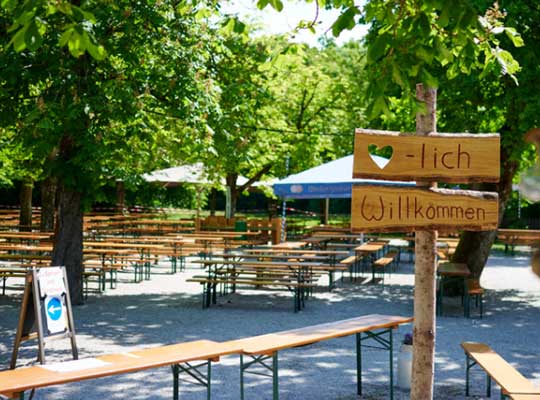 Ansicht des Außenbereichs des Waldheim Biergartens mit großen grünenden Bäumen an einem sonnigen Tag, im Vordergrund ist ein Willkommensschild zu sehen