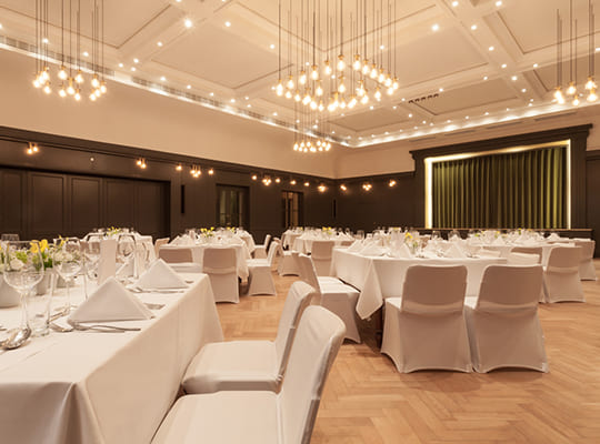 Nobel gestalteter Speisesaal mit braunen Wandeinheiten und wundervoller weißer Decke inkl. schönen Deckenleuchten. Vor der großen Bühne sind jede Menge Tische in weißer Farbe angerichtet.