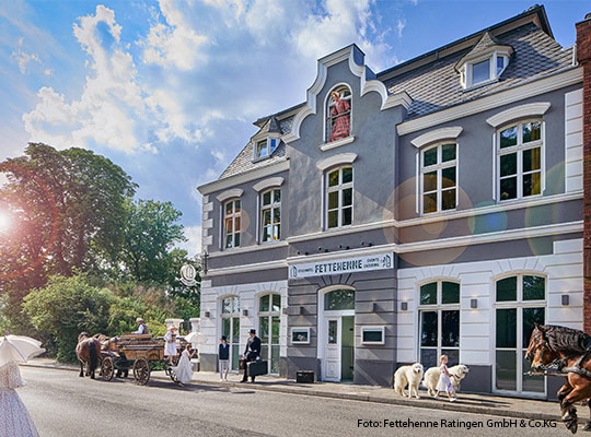 Ein großes gasthaus steht an einer Straße. Auf der Straße gehen Menschen im Sonnenschein spazieren. 