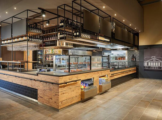 Ansicht einer sehr modern gestalteten Theke mit Bar, oben sind Weinflaschenhalter in Industrioptik und die Bar ist aus Holz 