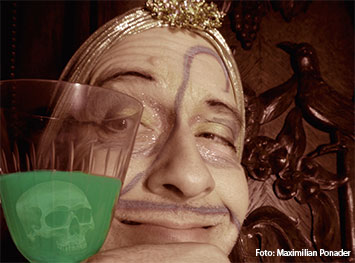 Ein lächelnder geschminkter Mann hinter einen Glas mit grüner Flüssigkeit mit einem Totenkopf drin