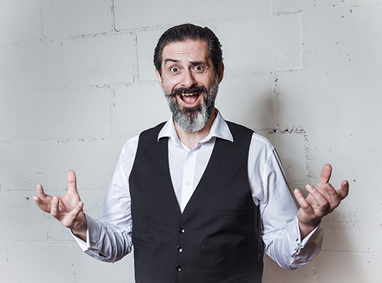 Carlos Coelho, der Schauspieler, steht vor einer gräulichen Wand, hält seine Hände nach oben und zieht dabei eine witzige Grimasse. 
