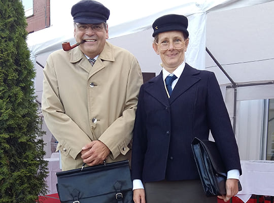 Ein Schauspieler und eine Schauspielerin stehen nebeneinander mit jeweils einer Aktentasche. Der Mann hat zudem eine Pfeife im Mund.