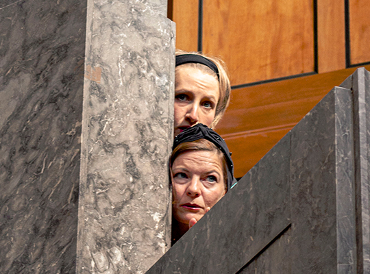 Zwei Schauspielerinnen beobachten das Geschehen versteckt hinter einer Mauer.