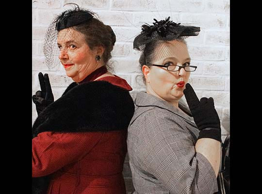 Eine Frau in rot und eine in grau posieren mit einer zur Pistole geformten Hand.