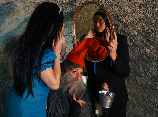 Eine Frau im blauen Kleid schaut in einen Spiegel, vor ihr steht ein Zwerg mit roter Mütze.