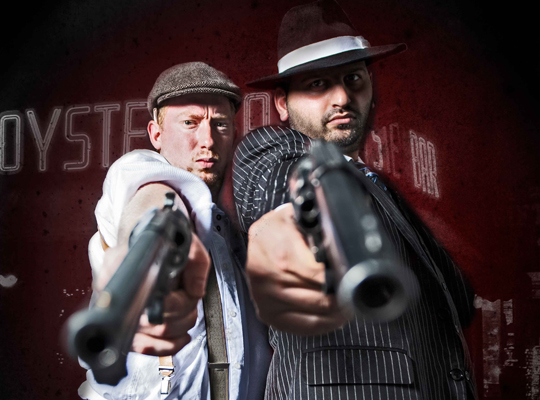 Zwei Schauspieler, verkleidet als Mafiosi bedrohen den Zuschauer mit Pistolen.