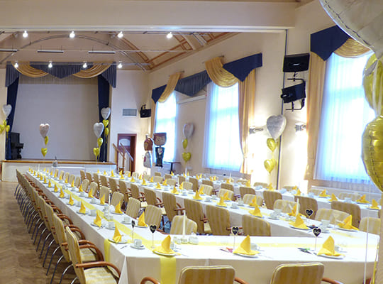 Großer Festsaal mit Bühne, in weißgoldenen Farben dekoriert und großen Tischtafeln.