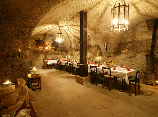 Gewölbekeller aus dem 14. Jahrhundert mit großem Holzbalken in der Mitte, altem Kaminofen langer Tischtafel und Tierfellen an der Wand.