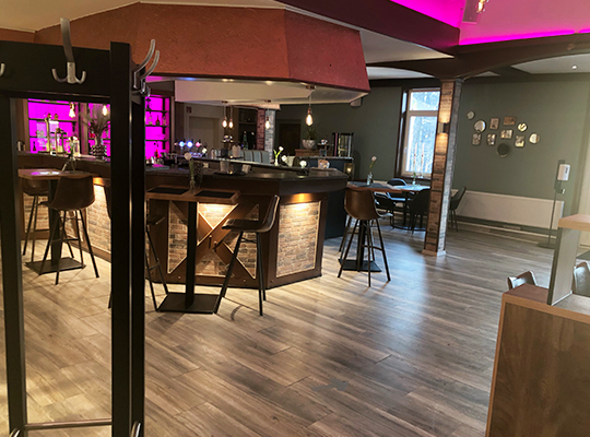 Modern gestalteter Barbereich mit bequemen Barhockern, einer Garderobe im Vordergrund und neon-rosa beleuchteter Barwand sowie Decke.