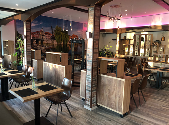 Viele bequeme Sitzgelegenheiten im Restaurantbereich mit modernen Raumtrennern, heller beleuchtung durch die Fenster und Lampen sowie ein großes Wandbild von Lüneburg.