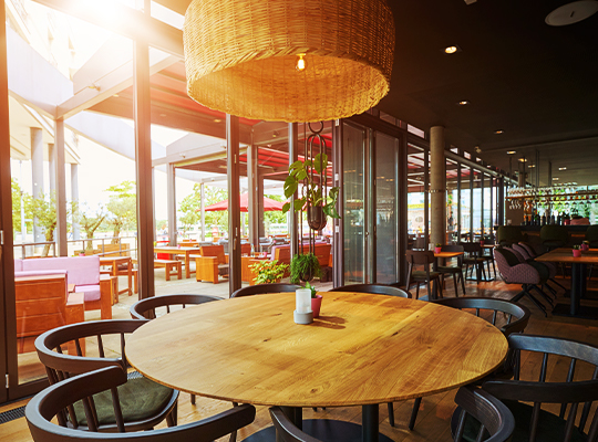 Hellbeleuchteter Restaurant-Innenraum des Beviamo Mainz, mit Blick auf die Terrasse sowie den Bar-Bereich. Im Vordergrund ein großer runder Tisch mit einer riesigen aus Holz gewebten Lampe.