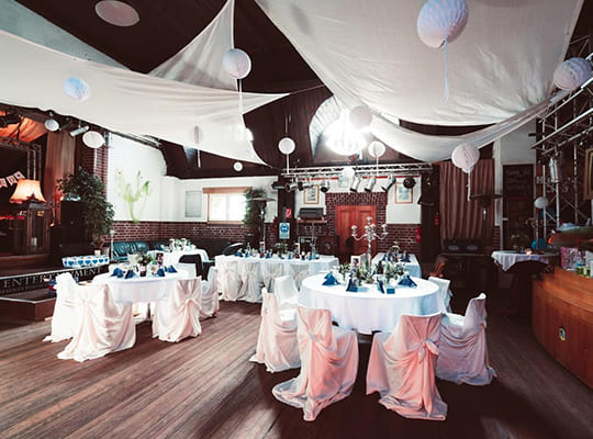 Großer Festsaal mit Bühne, Theke und Parkettboden. In der Mitte des Saals befinden sich Tische und Stühle mit weißen Hussen. Von der Decke hängen weiße Decken.