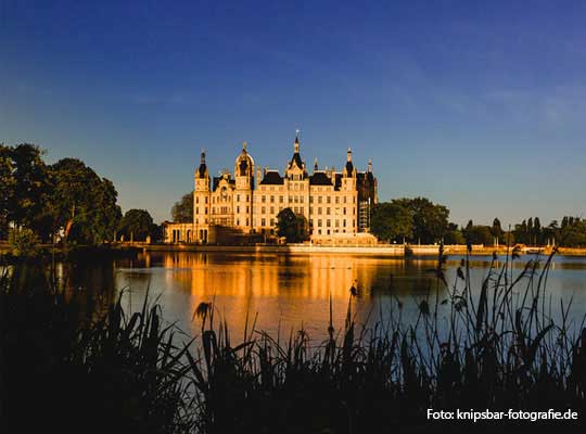 See mit Schloss im Hintergrund, Schloss wird von Sonnenlicht angestrahlt, strahlend blauer Himmel