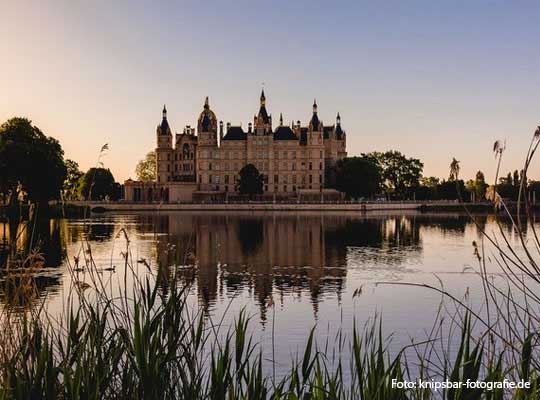 Schloss in Abenddämmerung mit See im Vordergrund, Gräser umspielen das Bild