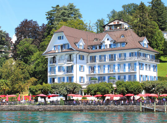 Blauer See im Vordergrund und schönes weißes Haus im Grünen, rote Sonnenschirme vor dem See