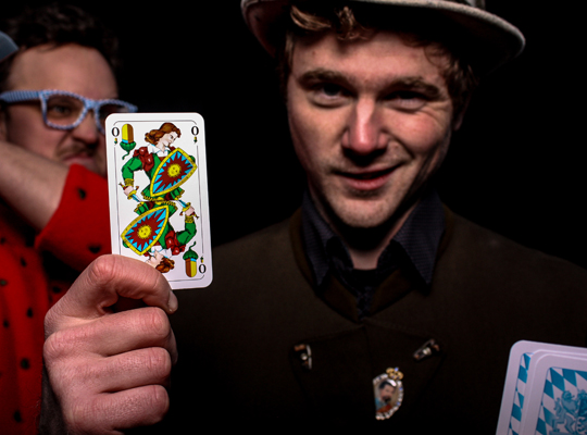 Mann mit Spielkarte in der Hand wird von hinten von einem anderen Mann mit Messer angegriffen - Krimidinner Schafkopfkrimi