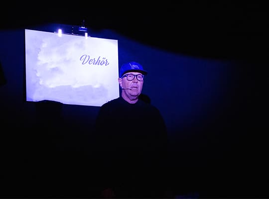 Schauspieler im Dunkel mit einer hellen Tafel hinter sich, mit der Aufschrift Verhör.