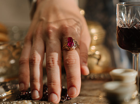 Hand mit einem Diamantring greift nach Schmuck, daneben steht ein edler Weinglas gefüllt mit Rotwein.